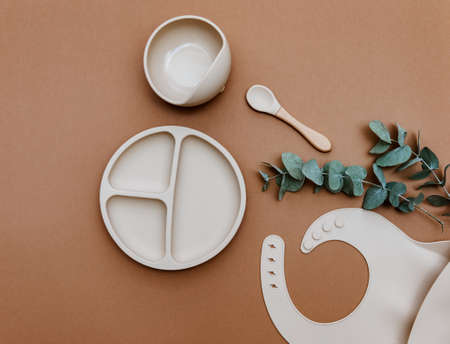 Baby food accessory on brown background. Neutral silicone bib and bowl. Kids dishware kit, serving baby foodの写真素材