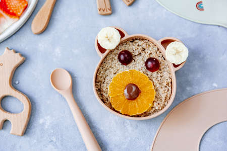 Funny bowl with oat porridge with bear faces made of fruits and berries on blue background. Food for kids idea, top view, copy space.の写真素材