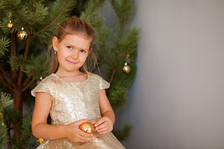 Portrait of little cute smiling girl in golden dress with christmas tree on background holding a golden Christmas tree toyの写真素材