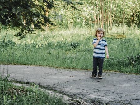 curly-haired boy in a striped T-shirt stands on the asphalt path pensive and worried and surprised,...