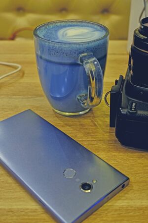 Trendy drink: Blue latte. Top view of hot butterfly pea latte tea or blue spirulina latte and photo camera nearby on wood table background.の写真素材