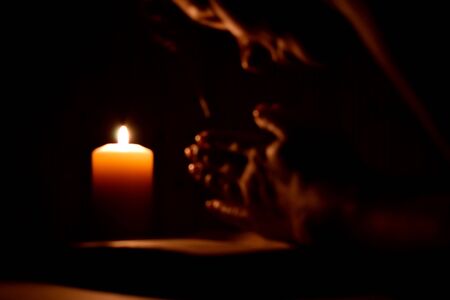 Religious prayer with a candle in complete darkness. Request for help from God. Praying in bible. Palms on old book on black background.の写真素材