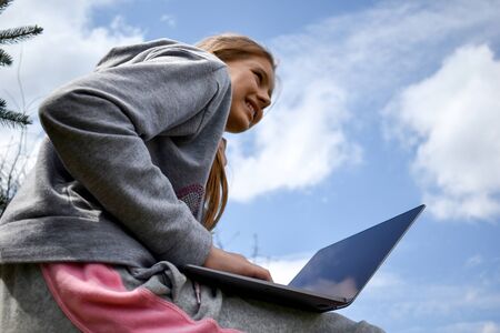 Girl with laptop on green grass. Remote schooling. The child works at the computer