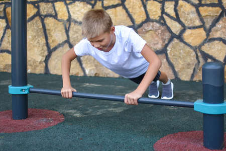 boy trains on the outdoor sports simulator. Sports child in the summer on playground. gymnastic exercises outside. boy overcoming efforts. achieve a difficult goal.の写真素材