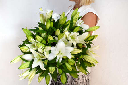 Woman with a bouquet of white lilies. Beautiful fresh delicate flowers for the holiday. Woman with beautiful flowers in her hands indoors background. Bouquet of lilies for a flower shop.の写真素材