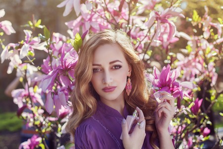 Outdoor portrait of a young beautiful lady using perfume near magnolia tree with flowers. Model looking at camera. Girl wearing stylish clothes. Female spring fashionの写真素材