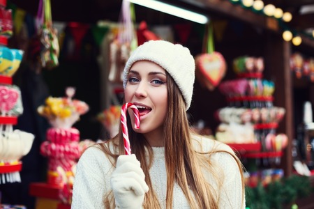 Street portrait of a smiling beautiful young woman biting Christmas candy cane and looking at camera. Lady wearing classic winter knitted clothes. Festive Christmas fair as background. Close up.の写真素材