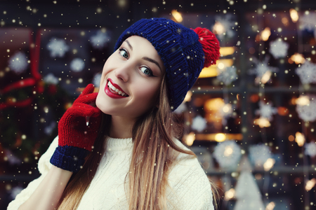 Street portrait of smiling beautiful young woman wearing classic winter knitted clothes. Model looking at camera. Festive garland lights. Magic snowfall effect. Close up. Toned.の写真素材