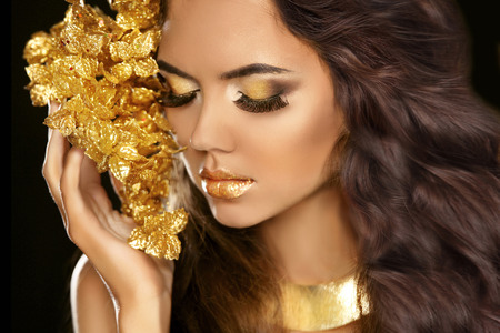 Golden makeup eyes closeup. Beautiful young woman in gold with flowers isolated on black background. Long brown hair.の写真素材