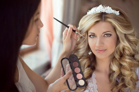 Professional Stylist makes makeup bride on the wedding day. Beautiful smiling blond woman with long curly hair style.の写真素材