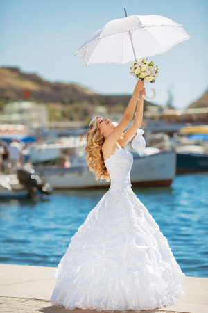 Beautiful smiling bride girl in wedding dress with white umbrella and bouquet of flowers walking on seafront, outdoors portraitの写真素材