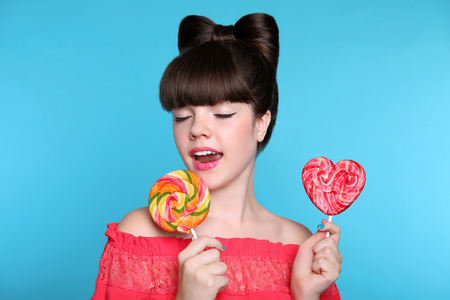 Lollypop. Beauty fashion teen girl with colourful eating colourful lollipop. Happy funny teenage with bow hairstyle solated on studio blue background. Sweets.の写真素材
