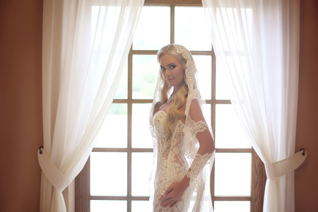 Elegant bride wedding portrait. Fashion white lace veil. Beautiful blond model posing by the window with curtain. Indoor bridal portrait. Makeup and Hairstyle.の写真素材