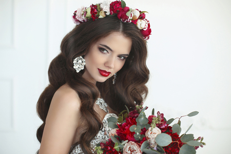 Makeup. Wedding Hairstyle. Beautiful bride brunette woman with bouquet of flowers, expensive jewelry close-up. Fashion portrait of elegant lady posing in white modern interior.の写真素材