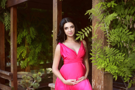 Pregnant woman posing in the park. Pregnancy, outdoors. Happy motherhood. Attractive young brunette in pink dress touching belly on nature spring countryside.の写真素材