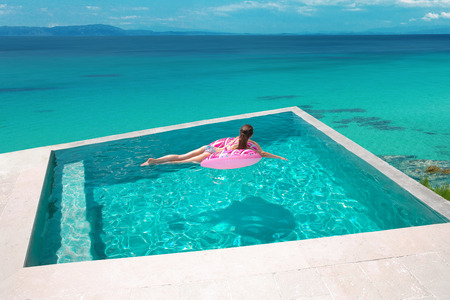 Sexy girl relaxing on inflatable pink donut in the infinity swimming pool at luxurious villa resort....