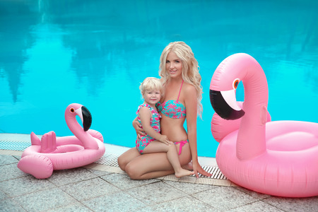 Summer family vacation. Fashion look blond girls portrait. Beautiful Mother holding her little daughter, wears in swim wear posing with Inflatable Flamingo Pool Float by swimming pool on resort.の写真素材