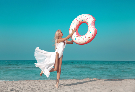 Summer Vacation. Beautiful blonde model in white swimwear jumping with inflatable donut float mattress on the beach. Wellness and beauty healthy concept.の写真素材