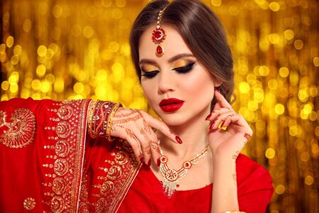 Portrait of beautiful indian girl in red bridal sari over golden bokeh. Young hindu woman model with kundan jewelry. Traditional Indian costume lehenga choli. Henna painting, mehendi on bride's hands.の写真素材
