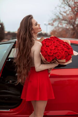 Happy smiling woman with rose bouquet of flowers posing by new red car. Beauty outdoor autumn portrait. Valentines day. Romantic expensive luxury gift.の写真素材