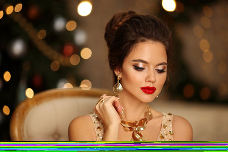 Luxurious portrait of elegant woman with wedding hairstyle and makeup. Beautiful brunette girl with golden jewelry in prom dress sitting on modern chair over bokeh lights xmas decorations.の写真素材