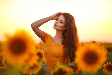 Beautiful young girl enjoying nature on the field of sunflowers at sunset. Summertime. Attractive brunette woman with long healthy hair.の写真素材