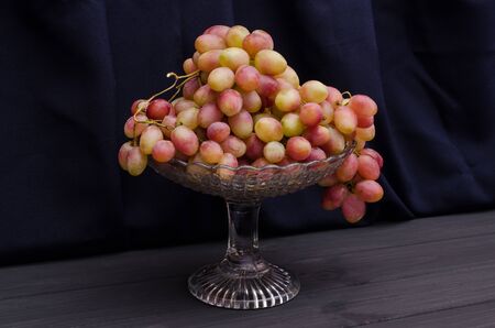 Red and pink grapes in a vase on a wooden table on a black fabric background. Fresh dessert, healthy...