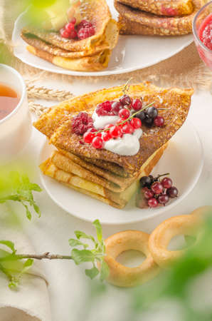 Pancakes with berries and sour cream for the Russian Maslenitsa holiday on a white background with bokeh of greensの写真素材
