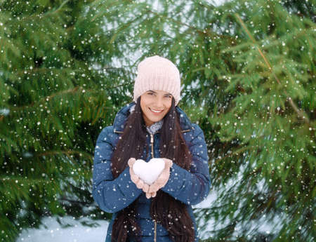 beautiful young woman holding a heart-shaped snowball in her hands, on a snowy winter day in the...