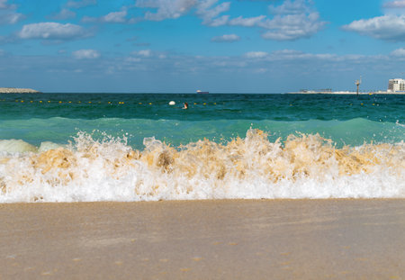 Gentle waves roll onto the sandy beach, sparkling in the sunlight. A vivid blue sea stretches to the horizon, under a bright sky filled with fluffy clouds.の写真素材