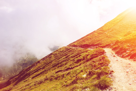 Toned serpentine path going down in the Carpatian mountains and a big fog cloud at the background, Bucegi natural park, Romania, on sunny summer day.の写真素材