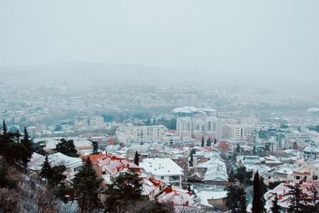 TBILISI, GEORGIA - JANUARY 1, 2016: A panoramic view to Tbilisi city center cowered with snow from Mtatsminda mountain on cloudy foggy winter morning with trees at the foreground.のeditorial素材
