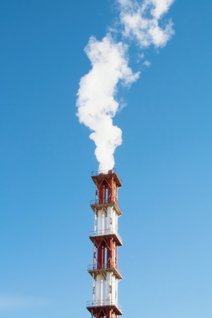 Red and white industrial chimney with a cloud of white smoke against blue clear sky. の写真素材