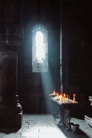 A dark background of old Armenian christian church monastery interior with a sun ray falling to the candles from a window. Abstract christian religion belief and travel concept.のeditorial素材