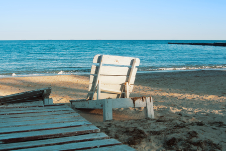 One lonely old white vintage wooden sun lounger standing at the abandoned beach after the end of a summen season in the evening sunlight. の写真素材