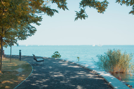 A view of Balaton lake with white yachts at the horizon and trees, bench and footpath at the foreground on sunny autumn day, Tihany, Hungary.の写真素材