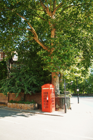 LONDON, ENGLAND - AUGUST 01, 2013: Bright red classical english phone booth on a crossroad in the park under a big tree on sunny summer day.のeditorial素材