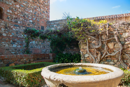 MALAGA, SPAIN - FEBRUARY 16, 2014: A courtyard with old stone brick walls, an old medieval stone fountain and plants at the gardens of the famous Palace Fortress of Alcazaba in Malaga, Andalusia.のeditorial素材