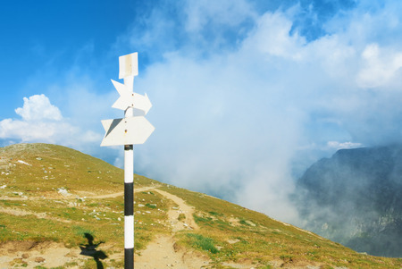 A signpost direction white sign arrow near the tracking path at the peak top of the mountain showing...