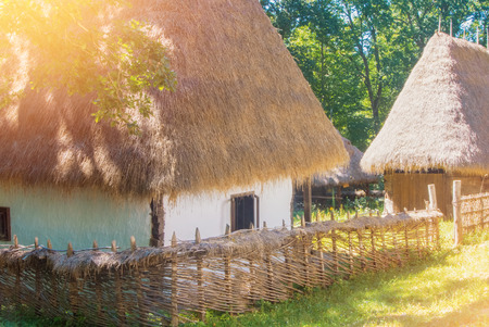 Old beautiful traditional romanian ukranian farm village house with white walls, roof covered with straw, wooden fence wickered by hand and a garden with wooden barn in warm sunlight on summer day.の写真素材
