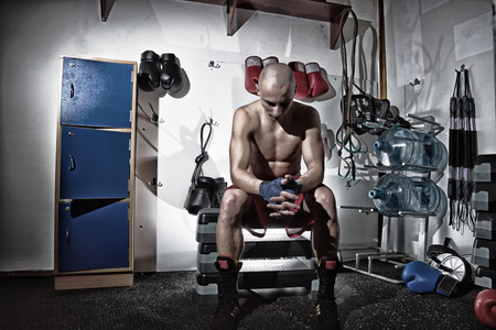 Male boxer full lenght portrait sitting in a changing room, looking down, holding hands, bare chested with a sport equipment behind himの写真素材