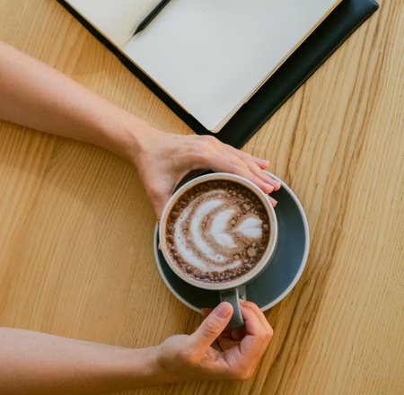 Cozy workspace aesthetic with hands holding cocoa and open notebook.Close-up of hands holding cocoa beside open notebook in workspaceの写真素材