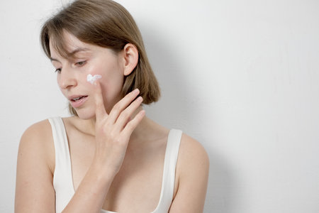 Young woman applying cream on her face against white background. Skin care conceptの写真素材