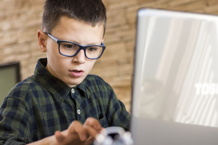 Boy With Glasses Using Laptop Computer While Sitting on Desk at Home. Studying Concept.
