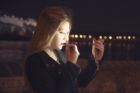 Young Beautiful Woman Putting Makeup and Looking at Himself on Smart Phone. Glow Bokeh Night City Lights in the Background.の写真素材