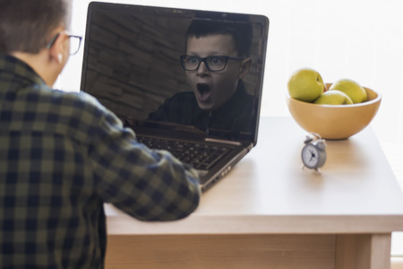 Shocked Boy With Glasses Using Laptop Computer While Sitting on Desk at Home. Expressive Face in Reflection.の写真素材