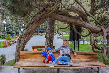 A girl with a child on a bench under the branches of a tree in Borjomi Park, on a rainy spring day. Georgia 2019.の写真素材
