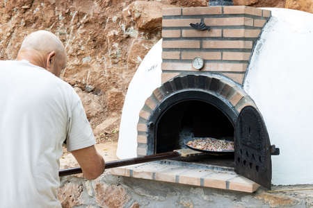 Man putting pizza in handmade white painted wood oven built outdoors with shovelの写真素材
