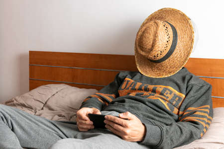 Man with headphones, mobile phone and face mask, sitting on his bed and confined to his roomの写真素材