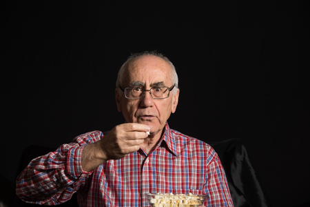 old man, wearing checked shirt eats popcorn and watches televisionの写真素材